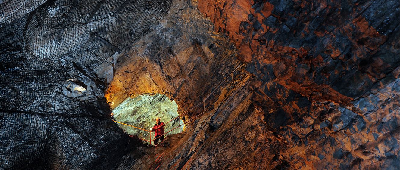 A photograph of Dudley Caverns with a person in a red jacket and yellow hard hat standing behind a fenced path.