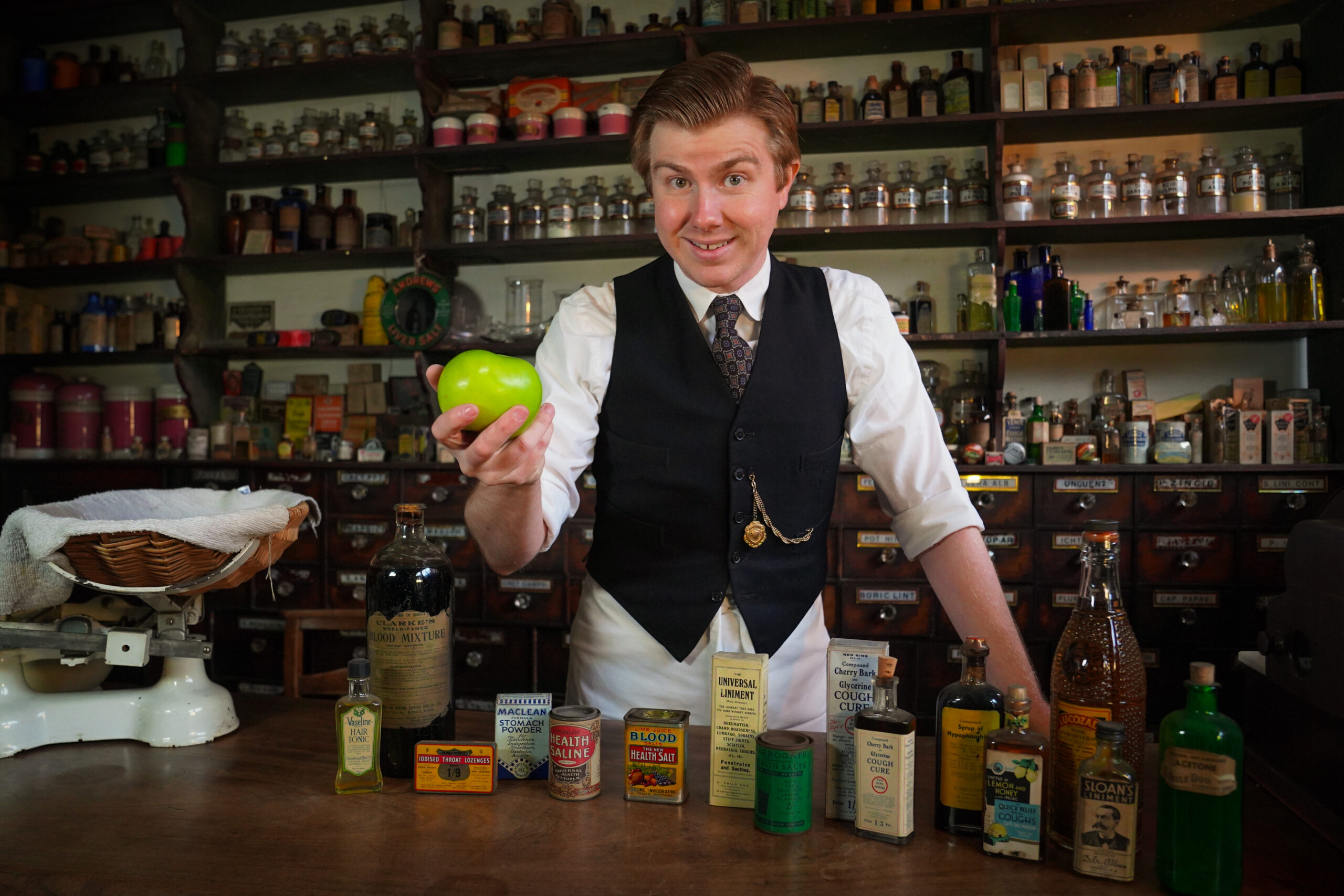A historic character holds a green apple in the chemist.